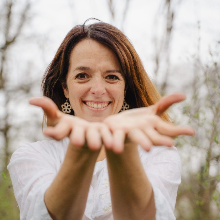 Frau mit braunen Haaren, die lächelt und beide Hände mit offenen Handflächen ausstreckt.